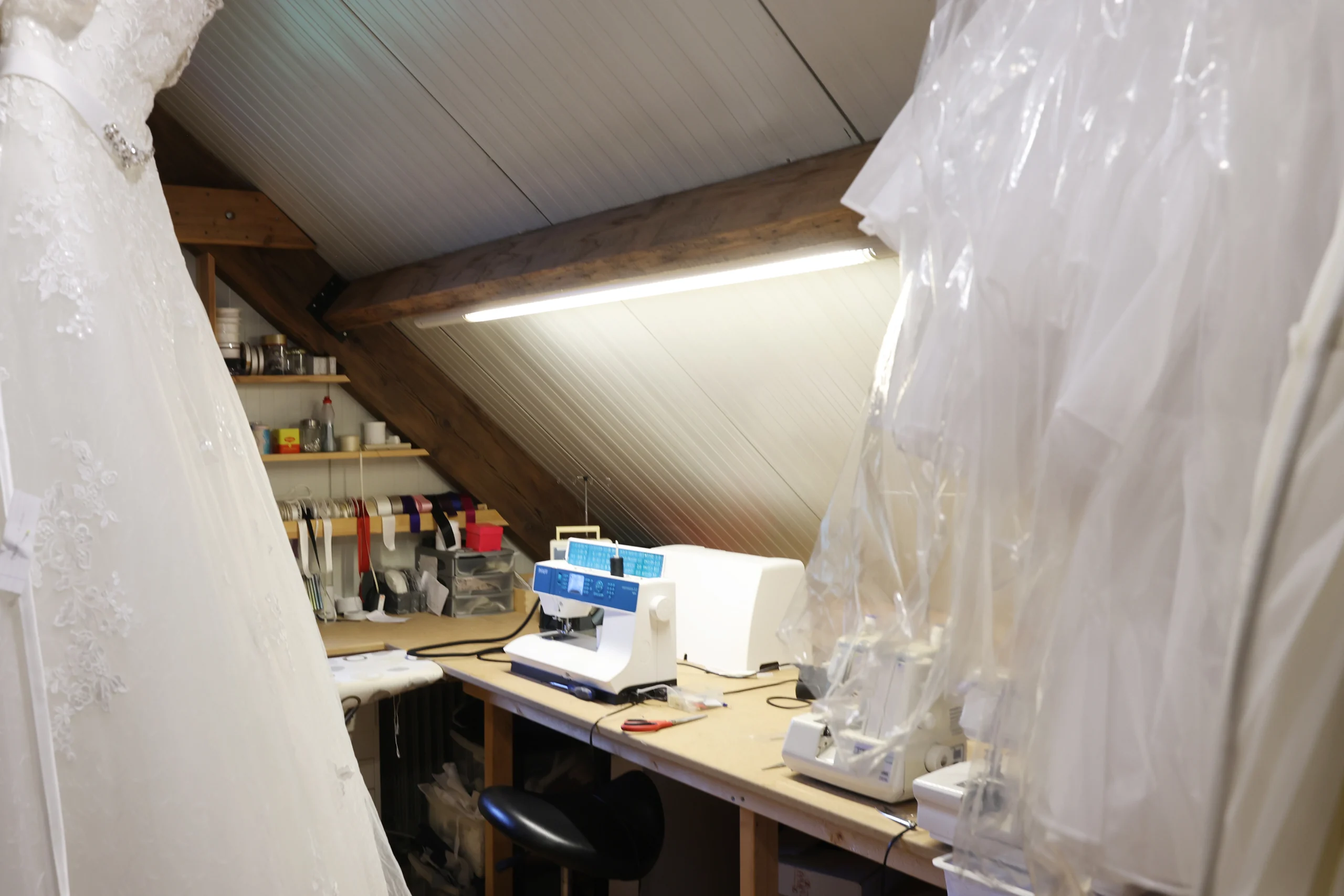 Dit is de naaihoek in de bruidsboetiek met een naaimachine op een werktafel en prachtige bruidsjurken hangend in beschermhoezen.